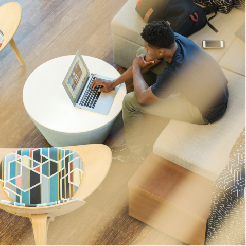 A birds eye view of a young man. He is a Black man sitting in a public work space wearing a blue jumper and beige trousers. He is working on a silver laptop which is sitting on a round white table.