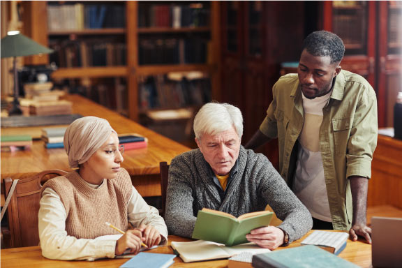 Two men and a woman looking at a book with a green cover together in a library. The man in the middle is holding the book. He is a white man with grey hair and wearing a grey jumper. To his left there is a young woman of south Asian ethnicity sitting down wearing a beige head scarf and jumper. On the far right there is a Black man standing up wearing a khaki green shirt.